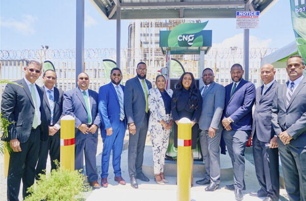 NGC | A group of eleven people in business attire stand together in front of a CNG station under a canopy. Barbed wire fencing, signage, and industrial structures are visible in the background.