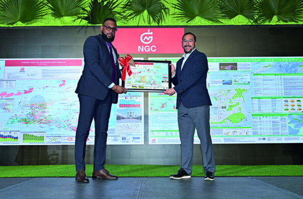 NGC | Two men in suits stand on a stage, holding a framed certificate together. A large display with maps and charts is visible in the background beneath a green “NGC” logo.