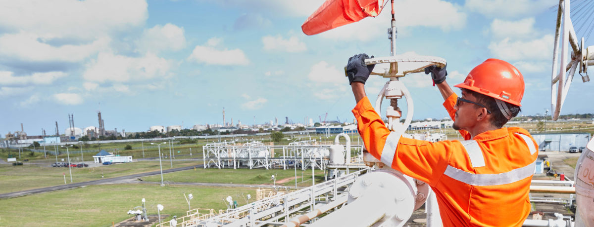 NGC | Under a partly cloudy sky, a worker in orange safety gear diligently operates a valve at an industrial site, showcasing the company's commitment to safety and precision—key aspects highlighted in the company profile.