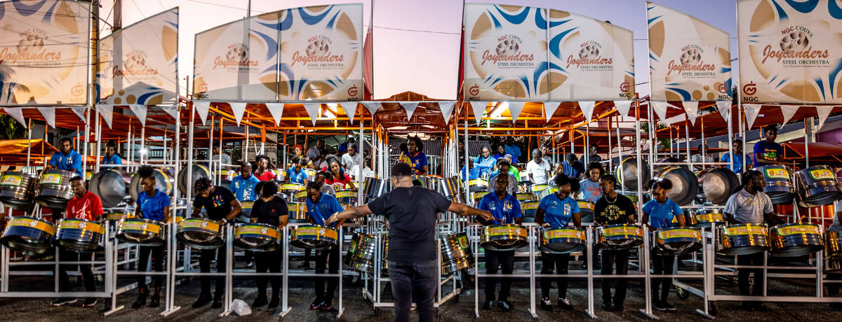 NGC | A conductor leads a large steel pan orchestra, with musicians in blue shirts playing at an outdoor event under branded banners, embodying the spirit of corporate social responsibility.