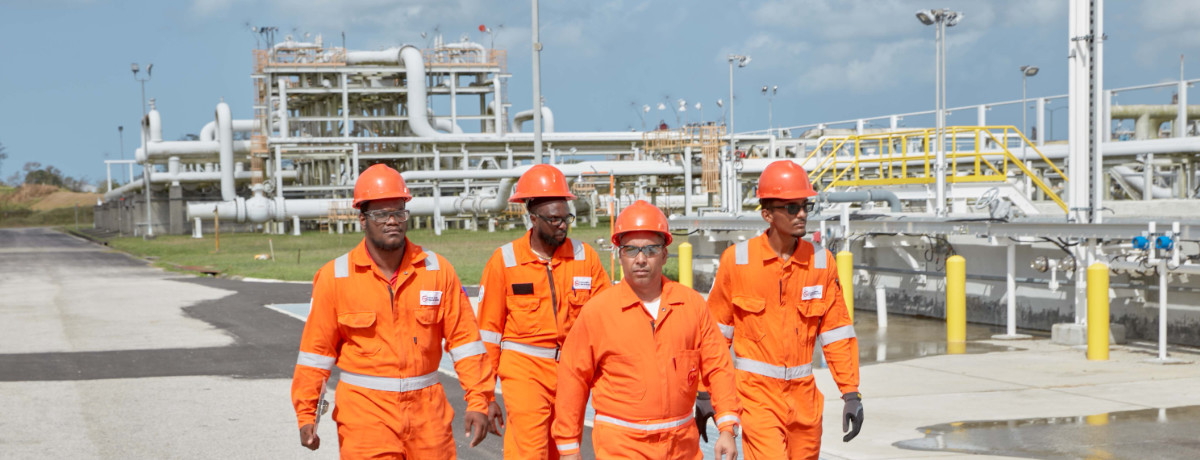 NGC | Four workers in orange uniforms and hard hats walk at an industrial plant, navigating the labyrinth of pipelines and equipment, mindful of their tight schedule under a cloudy sky.