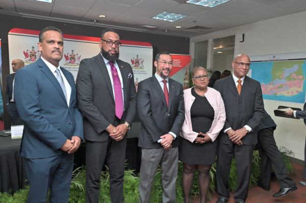 NGC | Five people in formal attire stand side by side in an office setting with a backdrop featuring logos.
