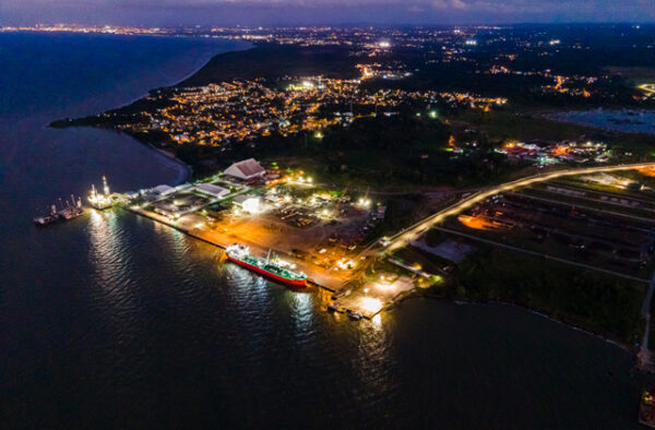 NGC | Aerial view of a port at night with a docked cargo ship, illuminated facilities, and lights from nearby town visible in the background.