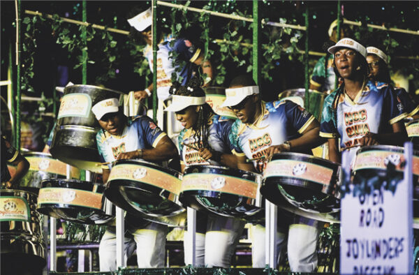 NGC | A group of musicians play steel pan drums on stage, wearing matching uniforms and caps, surrounded by green decorations.